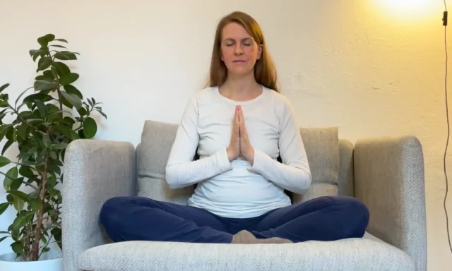 Die Yogalehrerin Anna Sophie Pietsch sitzt im Schneidersitz auf einem Sessel und hat die Hände im Anjali Mudra.